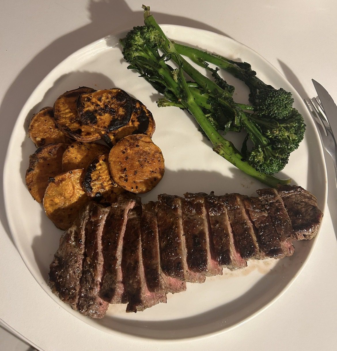 Overhead plate of sliced sirloin steak with charred sweet potato and broccolini on a dark board